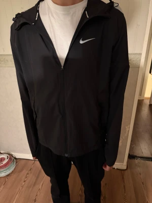 Svart vindjacka från Nike med huva - Svart vindjacka från Nike med klassisk logga på bröstet. Jackan har hel dragkedja, justerbar huva och två sidofickor. Lätt och smidig modell i syntetmaterial, perfekt för blåsiga dagar. Enkel och stilren design som funkar till det mesta.