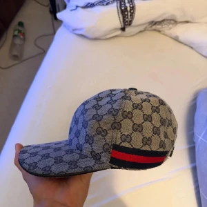 Gucci keps med GG-mönster, M - Säljer en snygg Gucci keps i beige och mörkgrått GG-monogram. Kepsen har klassisk böjd skärm, justerbart spänne bak i skinn och ikoniska röd-blå ränder på sidorna. Tillverkad i bomullscanvas med detaljer i skinn. Perfekt för dig som vill sticka ut med lyxig streetstyle.