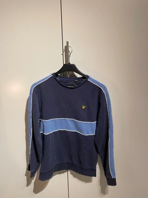 Blå sweatshirt från Lyle & Scott - Snygg marinblå sweatshirt från Lyle & Scott i storlek M. Tröjan har ljusblå paneler och vita detaljer längs ärmar och bröst, samt den klassiska gula logon på vänster sida. Rund hals och långa ärmar. Priset är diskuterandet vid snabb affär!