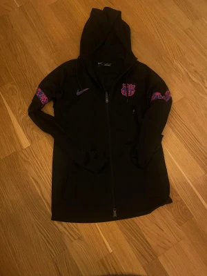 Svart SÄLLSYNT! Nike FC Barcelona hoodiejacka - Svart hoodiejacka från Nike med FC Barcelona-logga på bröstet och lila detaljer på ärmarna. Jackan har hel dragkedja, huva med snörning och är tillverkad i Dri-FIT polyester. Perfekt för dig som älskar fotboll och vill ha en sportig look. OBS DENNA VARA SÄLJS INTE OFFENLIGT LÄNGRE.