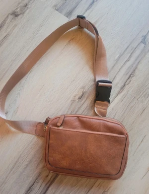 Brun axelväska i skinn - Snygg brun axelväska i skinn med justerbart band och praktisk dragkedja. Väskan har en enkel, rektangulär form och en framficka för småsaker. Perfekt för att bära med sig det viktigaste och passar till en chill streetstyle.