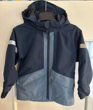 Blå vindjacka från Everest med huva - Snygg tvåfärgad blå vindjacka från Everest med huva och reflexdetaljer på ärmar och rygg. Jackan har dragkedja framtill, justerbara ärmslut med kardborre och praktiska fickor. Perfekt för blåsiga dagar och utomhusaktiviteter.