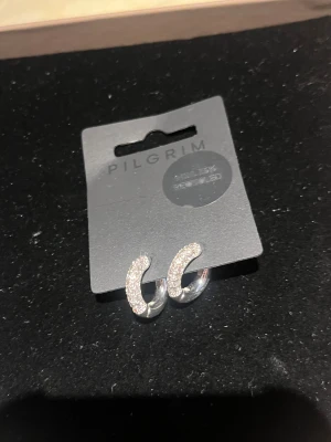 Glittriga silverörhängen från Pilgrim - Snygga örhängen i silverfärg från Pilgrim, designade som små hoops med glittriga stenar på framsidan. De är tillverkade av minst 75% återvunnet material och har en modern, stilren look som lyfter din outfit. Perfekta för dig som gillar bling och vill sticka ut. Dessa är även inte använda.