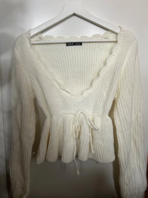 Vit stickad topp/tröja från SHEIN - Supersöt vit stickad kofta från SHEIN med v-ringad hals och volangkant. Koftan har långa ärmar, ribbad struktur och knytband i midjan som ger en snygg siluett. Perfekt för att addera en gullig touch till din outfit.