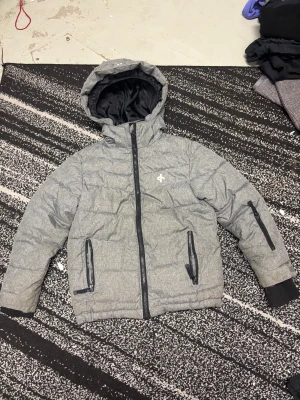 Grå pufferjacka från Cross Sportswear - Snygg grå pufferjacka med huva från Cross Sportswear. Jackan har svarta detaljer, flera dragkedjor och quiltad design. Perfekt för kalla dagar med sin varma och fluffiga känsla. Fodrad insida och ribbade muddar vid ärmslut. Stilren logga på bröstet.