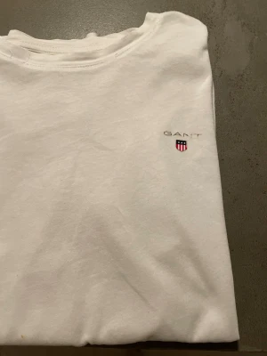 Vit t-shirt från GANT - Vit t-shirt från GANT med klassisk rund hals och korta ärmar. På bröstet finns en liten broderad GANT-logga och ett emblem i rött, blått och vitt. T-shirten är gjord i mjuk bomull och har en clean, enkel stil som funkar till allt.