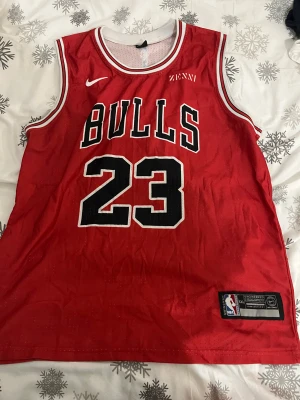 Chicago Bulls Jordan 23 baskettröja XXL - Säljer en röd Chicago Bulls baskettröja med Michael Jordans namn och nummer 23. Tröjan är ärmlös, har vita och svarta detaljer samt broderade loggor från Nike och NBA. Tillverkad i ventilerande mesh-material, perfekt för sport eller streetwear. Passar även XL bra.