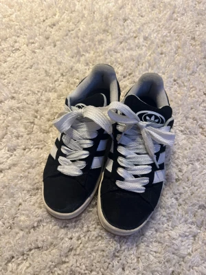 Adidas Campus svarta sneakers strl 40 - Svarta Adidas Campus sneakers med klassiska vita ränder och logga på plösen. Skorna har rund tå, platt sula och är tillverkade i mocka. Modellen är low cut. Perfekt för dig som gillar retrostil och streetwear.