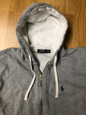 Grå hoodie från Polo Ralph Lauren - Grå hoodie från Polo Ralph Lauren med dragkedja och vit snörning i huvan. Tröjan har två fickor framtill och ett mörkblått broderat Polo-logo på bröstet. Mjuk insida och ribbade muddar vid ärmslut och nederkant. Perfekt för chill dagar. Pris är diskutrbarr mvh Rasmus! Perfekt julklapp🎁