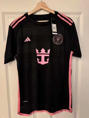 Inter Miami Messi #10 fotbollströja - Snygg svart Inter Miami-tröja från Adidas med rosa detaljer och Messi #10 på ryggen. Klubbmärke på bröstet, rosa axelränder och lätt, ventilerande polyester. Perfekt för att representera Messi och Miami.  Följ oss gärna på Instagram: @kickstoreuf