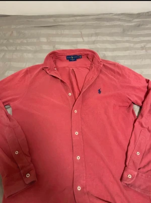 Röd skjorta från Ralph Lauren - Säljer en snygg rosa skjorta från Ralph Lauren i featherweight mesh. Skjortan har klassisk krage, långa ärmar och knappar framtill. Broderad blå logga på bröstet. Tillverkad i 100% bomull för en skön och luftig känsla.