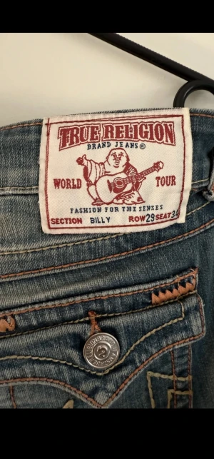 True Religion Billy jeans, blå, W29 - Säljer ett par True Religion Billy jeans i klassisk blå tvätt med snygga slitningar och kontrastsömmar. Jeansen har ikoniska bakfickor med lock och logga, samt silverfärgade knappar. Tillverkade i USA och i klassisk jeanskvalitet. Perfekta för dig som gillar street och premium denim.