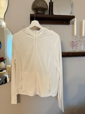Vit vindjacka med huva - Säljer en stilren vit vindjacka med huva och hel dragkedja framtill. Jackan har två fickor med dragkedja och ventilerande paneler på sidorna. Långärmad och lätt material som passar perfekt för aktiva dagar utomhus.