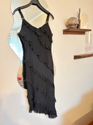 Svart volangklänning med smala axelband - Säljer en svart klänning med smala axelband och flera diagonala volanger som ger en cool och lekfull look. Klänningen har asymmetrisk nederkant och är tillverkad i ett lätt, flödande material som känns skönt mot huden. Perfekt för dig som gillar att sticka ut med detaljer.