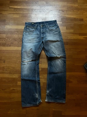 Blå slitna jeans från Lee - Säljer ett par blå jeans från Lee med snygga slitningar och hål på knäna samt fransiga benslut. Klassisk femficksmodell med normal passform och raka ben. Jeansen har en cool tvättad look och Lee-märke på bakfickan.