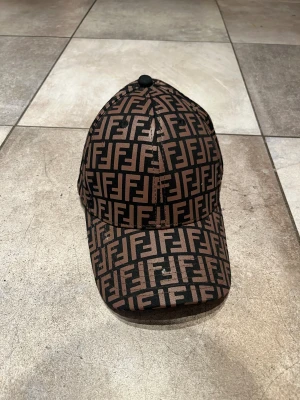 Fendi FF-logga keps brun/svart - Snygg keps från Fendi med ikoniskt FF-logga-mönster i brunt och svart över hela kepsen. Justerbar rem baktill med metallspänne och Fendi-tryck på insidan. Perfekt för dig som gillar lyxiga streetwear-accessoarer.