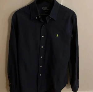 Mörkblå skjorta från Polo Ralph Lauren - Fin  mörkblå skjorta från Polo Ralph Lauren med  krage. Säljer då jag växt ur den. Köpte den i våras. Bra skick inget är sönder eller defekt med den. Hör av er vid eventuella frågor. MVH Måns 
