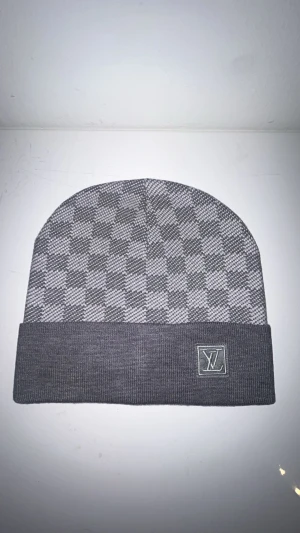 Grå rutig mössa med uppvik - Snygg grå mössa med uppvik och rutigt mönster. Mössan har en stilren design med diskret etikett framtill. Perfekt för dig som gillar enkel men trendig stil. Materialet känns mjukt och bekvämt mot huvudet.