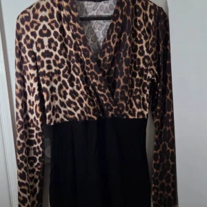 Leopardmönstrad omlottklänning - Snygg kort klänning med leopardmönstrad överdel och svart kjoldel. Omlott-skärning upptill och långa ärmar. Perfekt för dig som vill sticka ut med ett djurmönster och gillar trendiga detaljer.