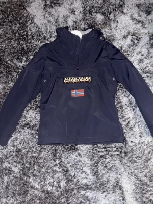 Mörkblå vindjacka från Napapijri - Snygg mörkblå vindjacka från Napapijri med huva och halv dragkedja. Jackan har en stor ficka framtill med Napapijri-logga och norsk flagga, samt justerbar huva med snörning. Perfekt för blåsiga dagar och streetwear-stil.