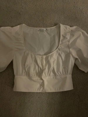 Vit blus med puffärm från NA-KD - Säljer en vit blus från NA-KD i storlek 38. Blusen har puffärmar, rundad halsringning och knappar framtill. Croppad modell med rynkade detaljer vid bröstet och midjan. Tillverkad i bomull för en fräsch och lätt känsla.