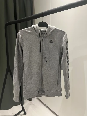 Grå hoodie från Adidas med tryck - Grå hoodie från Adidas med svart logga på bröstet och stort Adidas-tryck längs ena ärmen. Tröjan har dragkedja framtill, huva med svarta snören som är slitna (kolla bild).