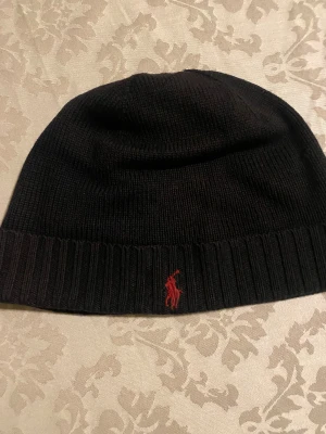 Svart stickad mössa från Polo Ralph Lauren - Svart ribbstickad mössa från Polo Ralph Lauren med klassisk röd broderad logga framtill. Mjuk och varm, perfekt för kyliga dagar. Enkel design med uppvikt kant och diskret branding som ger en clean look.