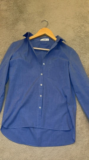 Blå skjorta från Mango Casual - Snygg blå skjorta från Mango Casual i storlek S. Skjortan har klassisk krage, lång ärm och en bröstficka. Tillverkad i mjuk bomull med knappar framtill och något längre bakstycke för en cool look.