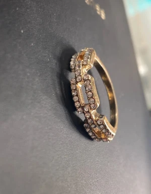 Guldfärgad ring med glittriga stenar - Snygg guldfärgad ring med unikt geometriskt mönster och massor av glittrande stenar runt hela designen. Perfekt för dig som gillar att sticka ut med accessoarer som verkligen syns. Ringen har en cool och modern vibe. Vet inte storleken