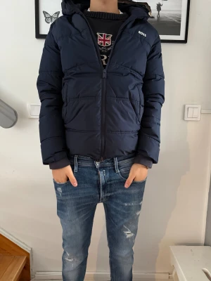 Mörkblå pufferjacka från BOSS - Snygg mörkblå pufferjacka från BOSS med huva och dragkedja framtill. Jackan har quiltad design och logga på bröstet. Perfekt för kalla dagar och har en clean, modern look som funkar till jeans och hoodie.