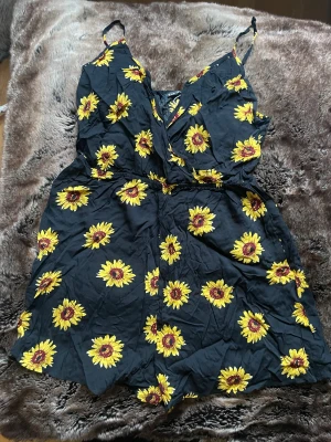 Svart playsuit med solrosmönster - Säljer en svart playsuit med färgglada gula solrosor över hela tyget. Playsuit har smala axelband och en v-ringad omlottfront med resår i midjan för en skön passform. Perfekt för dig som gillar blommiga prints och vill sticka ut.