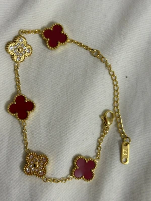 Guldarmband med röda klöver och kristaller - Unikt armband i guldfärgad metall med fem fyrklöverformade länkar, tre i röd emalj och två dekorerade med gnistrande kristaller. Kedjan har en snygg karbinlås . Perfekt för dig som gillar detaljer och färg. 18 k Guldpläterad 