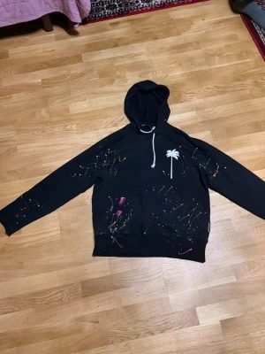 Svart hoodie Palm Angels med färgstänk - Svart hoodie från Palm Angels med vit palmtryck på bröstet och coola färgstänk i rosa, gul och blå över hela tröjan. Stor vit text på ryggen. Klassisk huva med snörning och magficka framtill. Perfekt statement-plagg för dig som gillar streetwear.
