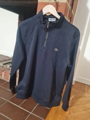 Marinblå half zip tröja från Lacoste - Snygg marinblå långärmad tröja från Lacoste med klassisk krokodillogga på bröstet. Tröjan har en ståkrage och half zip-dragkedja framtill. Regular fit och tillverkad i mjuk bomull, perfekt för chill dagar. Taggar kvar på plagget.