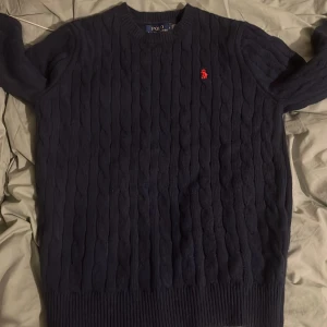 Mörkblå stickad tröja från Polo Ralph Lauren - Säljer en mörkblå stickad tröja från Polo Ralph Lauren med klassiskt kabelstickat mönster och röd broderad logga på bröstet. Tröjan har rund hals och långa ärmar, perfekt för dig som gillar stilren och tidlös design.