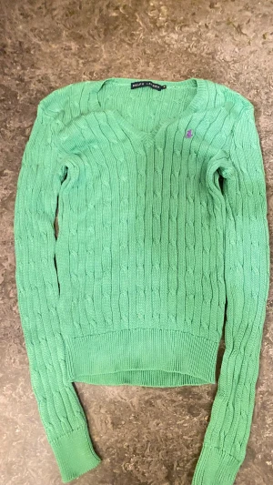 Grön stickad tröja från Ralph Lauren - Oanvänd Snygg grön stickad tröja från Ralph Lauren med klassiskt kabelmönster och lila broderad logga på bröstet. Tröjan har v-ringad hals, långa ärmar och ribbade muddar vid ärmslut och nederkant. Perfekt för dig som gillar klassisk stil!💕🗽( äkta såklart)