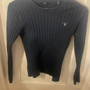 Mörkblå ribbad tröja från GANT - Snygg mörkblå ribbstickad tröja från GANT i storlek S. Tröjan har lång ärm, rund hals och ett litet broderat GANT-logo på bröstet. Materialet är mjukt och stretchigt, perfekt för dig som gillar klassisk och stilren look.