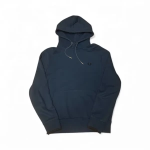 Marinblå hoodie från Fred Perry - Snygg marinblå hoodie från Fred Perry med klassisk logga broderad på bröstet. Tröjan har huva med snörning, ribbade muddar och en stor magficka. Perfekt för dig som gillar stilren och enkel design med premiumkänsla.