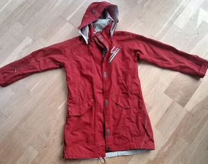 Röd regnjacka från Didriksons - Snygg röd regnjacka från Didriksons med huva, långa ärmar och praktiska fickor framtill. Jackan har dragkedja och knappar, samt vit logga på ryggen. Insidan är meshfodrad för extra komfort och ventilation. Perfekt för blöta dagar och outdoor.