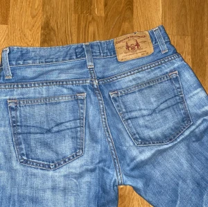 Blå bootcut jeans från Crocker Originals - Snygga blå jeans från Crocker Originals med klassisk bootcut-passform. Jeansen har fem fickor, bälteshällor och är tillverkade i 100% bomull. De har en ljus tvätt och raka sömmar, perfekt för dig som gillar retrovibbar och avslappnad stil.