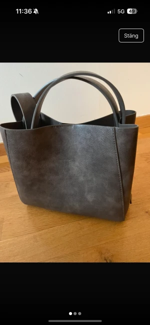 Mörkgrå stilren handväska i konstläder - Snygg mörkgrå handväska i konstläder med minimalistisk design. Väskan har både korta handtag och en längre axelrem samt ett innerfack med dragkedja för extra förvaring. Perfekt för dig som gillar enkel och modern stil.