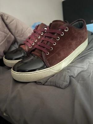 Lanvin sneakers i vinröd mocka - Lanvin sneakers med vinröd mocka och svarta lackade tår. Skorna har rund tå, snörning och vit platt sula. Snygg kontrast mellan det matta mockamaterialet och den blanka lacken framtill. Perfekt för dig som gillar lyxiga detaljer och stilren design. Snören, Dustbag och rengöringskit följder med. Boxen är slängd. Frakter idag 