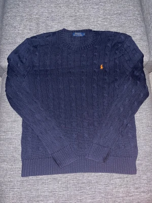 Marinblå stickad tröja Polo Ralph Lauren - Säljer en marinblå stickad tröja från Polo Ralph Lauren med klassiskt kabelstickat mönster och orange broderad logga på bröstet. Tröjan har rund hals, ribbade muddar och är långärmad. Perfekt för dig som gillar stilren och tidlös design.