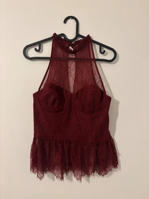 Vinröd spetskorsett med volang - Supersnygg vinröd korsett från zara med blommig spets, hög hals och volangkant nedtill. Toppen har formade kupor, transparent mesh upptill och dragkedja i sidan. Perfekt för dig som vill sticka ut med en chic och edgy look.