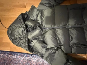 Mörkgrön pufferjacka från Everest - Säljer en mörkgrön pufferjacka från Everest med huva och grov dragkedja framtill. Jackan har en schysst quiltad look och är tillverkad i syntetmaterial. Perfekt för kalla dagar och har en clean, modern vibe. Helt ny pris kan diskuteras 