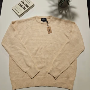 Beige stickad Stussy tröja M - Säljer en beige stickad tröja från Stussy i storlek M. Tröjan har ett stort grafiskt tryck i svart och rött på ryggen med Stussy-logga och figurer. Rund hals och ribbade muddar vid ärmslut och nederkant. Perfekt för dig som gillar streetwear och snygga detaljer.