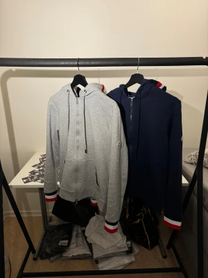 Moncler hoodie med dragkedja - Två hoodies från Moncler, en i ljusgrått och en i mörkblått. Båda har huva, hel dragkedja och långa ärmar med coola ränder i rött, vitt och blått vid mudden. Moncler-logga på ärmen och mjukt bomullsmaterial. Perfekt för chill eller streetwear.