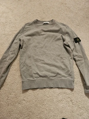 Grå Stone Island sweatshirt  - Grå sweatshirt från Stone Island med klassisk rund hals och långärmad passform. Tröjan har den ikoniska svarta patchen med gul broderad kompass på vänster ärm. Tillverkad i mjuk bomull för skön komfort och stilren look.
