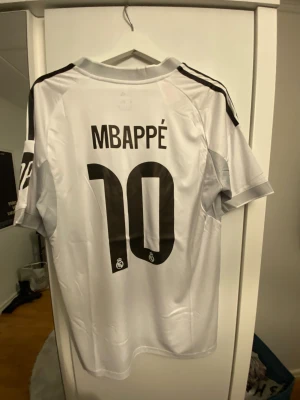 Real Madrid Mbappé 10 fotbollströja - Säljer en  Real Madrid fotbollströja från Adidas med Mbappé och nummer 10 på ryggen. Tröjan är vit med svarta detaljer, gula linjer och klubbmärke på bröstet. Materialet är lätt och andas, perfekt för match eller träning. Emirates Fly Better tryck på framsidan.