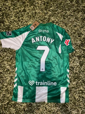  Real Betis 25/26 hemma tröja ANTONY 7 - Säljer en officiell Real Betis fotbollströja med gröna och vita vertikala ränder, tryckta sponsorer och klubbmärke på bröstet. Tröjan har korta ärmar, rund hals och namn samt nummer 'Antony 7' på ryggen. Materialet är lätt och andas, perfekt för match eller träning.